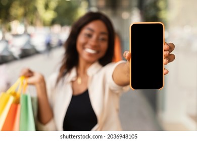 App For Online Store. Happy Black Woman Showing Smartphone With Blank Empty Screen Close Up To Camera And Holding Shopping Bags In Hand, Cheerful Lady Standing Outdoors Mall, Mock Up, Free Copy Space