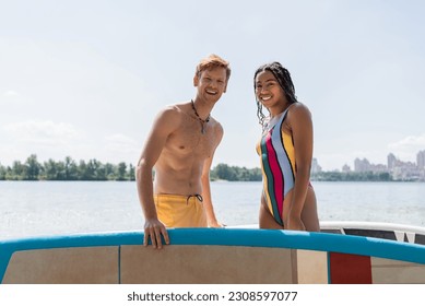 appealing african american woman in striped swimsuit and young redhead man in yellow swim shorts looking at camera near sup boards with lake and cityscape on background - Powered by Shutterstock