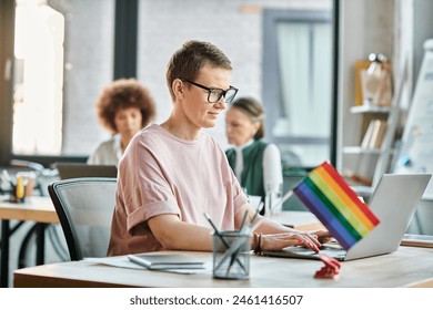 Appealing woman engrossed in work, with a laptop in front of her, with her diverse colleagues on backdrop, pride flag. - Powered by Shutterstock