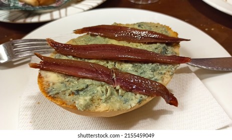 Appetizer Of Bread With Blue Cheese And Anchovies In A Typical Bar In Seville, In The South Of Andalusia (Spain). It Is On A White Plate With A Knife And Fork. There Is More Food On The Table.