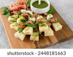 Appetizer with melon, mozzarella, prosciutto ham and fresh basil on stick with pesto sauce on a stone background. Finger food. Summer salad