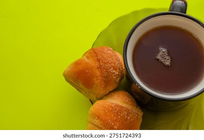 An appetizing cup of hot tea with surface bubbles served alongside golden homemade crescent rolls generously sprinkled with sugar. This minimalist still life on a vibrant lime green background creates a cozy atmosphere for breakfast or a pleasant coffee break.