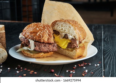 Appetizing And Juicy Half Cuted Burger With A Large Beef Patty And Cheddar Cheese Flowing From It, White Sauce, Pickled Cucumber And Onions On A White Plate With French Fries. Close Up