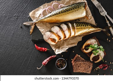 Appetizing smoked fish with spices, cutlery, pepper and bread on craft paper over dark stone background. Sandwich with smoked mackerel. Mediterranean food, herring fish, seafood, top view
