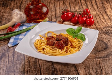 Appetizing spaghetti meatballs served on white plate, garnished with fresh basil. Rich red cherry tomatoes, garlic, chili, green onion, olive oil bottle complement rustic wooden background