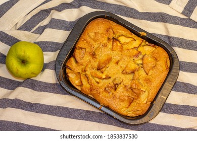 Apple сharlotte in the black cake tin and a green apple on the striped tablecloth. 