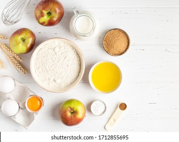 Apple pancakes or apple muffins ingredients top view. Flour, eggs in eggholder, bottle of milk, brown sugar, cinnamon powder, fresh apples, melted butter and baking powder. White wooden background. - Powered by Shutterstock