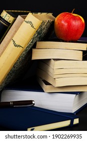 An apple with a stack of old, hardbound classic books, a notebook, and a fountain pen.
