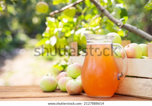 Apples, box of apples and pitcher of fresh\
apple juice on wooden table with garden background. Harvest of\
apples. Juice\
production.