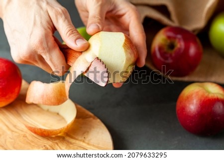 apples of different varieties are seen on dark stone and wooden background.  A woman is peeling the skin of an apple using a fruit knife. Healthy eating and cooking concept