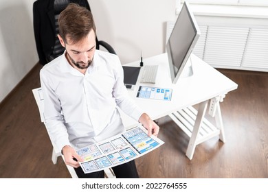 Application interface development. Man holds a sheet with app layouts. Application interface is depicted on paper. Web designer in his office. UX designer next to work table. Application design.
