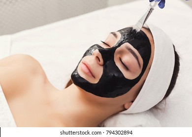 Apply Black Face Mask, Spa Beauty Treatment And Skincare. Woman Getting Facial Cleansing Mask By Beautician At Spa Salon, Closeup