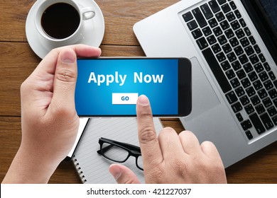 Apply Now Message On Hand Holding To Touch A Phone, Top View, Table Computer Coffee And Book