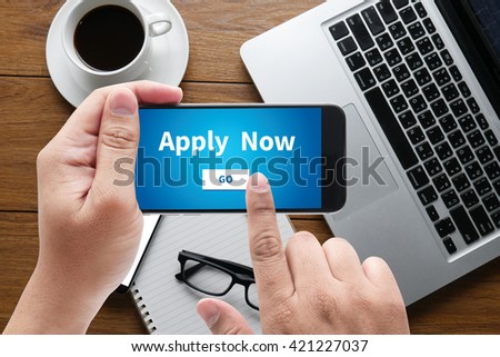 Apply Now message on hand holding to touch a phone, top view, table computer coffee and book