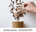 Apply now symbol. Concept word Apply now on wooden blocks. Beautiful white background with coffee cup. Businessman hand. Business and Apply now concept. Copy space