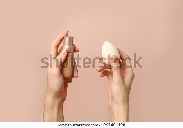 Applying foundation on\
makeup sponge. Woman\'s hands with neutral manicure holding bottle\
of concealer or toner foundation, cream and beauty blender, make up\
artist background
