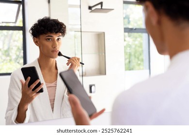 Applying makeup, transgender woman holding smartphone and looking in mirror. Beauty, cosmetics, self-care, grooming, transformation, confidence - Powered by Shutterstock