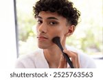 Applying makeup, young man using brush for contouring at home, focusing on face. Beauty, cosmetics, skincare, self-care, routine, mirror