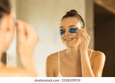 Applying under-eye patches, woman smiling at mirror in bathroom. Self-care, skincare, beauty, relaxation, facial, wellness - Powered by Shutterstock