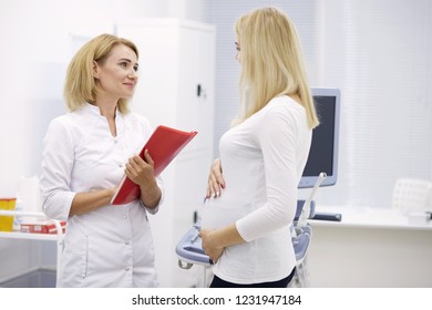 Appointment With The Doctor In The Clinic. Pregnant Woman Asking Questions Of A Experienced Doctor. Help, Advise. Side View Photo