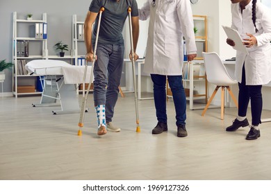 Appointment With Orthopedic Surgeon. Patient With Broken Leg Or Ankle Sprain Stands Up From Examination Bed And Walks On Crutches Assisted By Diverse Hospital Staff Of Professional Doctors And Nurses