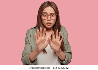 Apprehensive scared female stretches hands, refuses of something extreme, being terrified by awful proposal, asks not bother her, stares with bugged eyes through spectacles, stands indoor alone