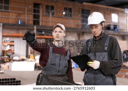 Apprentice gestures while supervisor holds clipboard at metal storage facility