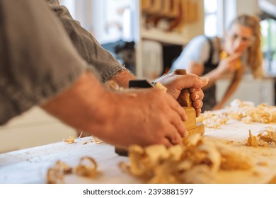 Apprentice observing with interest as a carpenter planes wood in a workshop