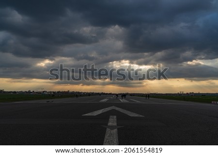 An approaching storm low over Tempelhofer Feld. The setting sun shines through the dark clouds. In the foreground a runway of the former Tempelhof Airport with an arrow straight ahead.