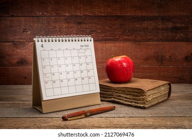 April 2022 - spiral desktop calendar with old book or journal and apple against rustic barn wood background, time and business concept
