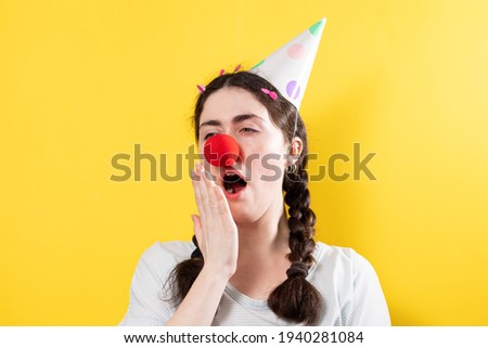 April's fool day concept. Portrait of a bored, yawning woman in a cap and with a red round false nose. Yellow background.