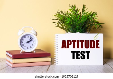 APTITUDE TEST is written in a notebook next to a green plant and a white alarm clock, which stands on colorful diaries.