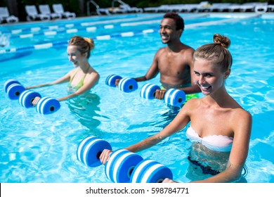 Aqua Fitness At Resort. Two Healthy Smiling Young  Women With Dumbbells And Their Male Instructor Standing At Swimming Pool And Doing Exercise On Summer Day Outdoors