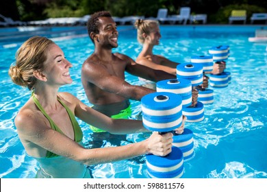 Aqua fitness at resort. Two healthy smiling young women with dumbbells and their male instructor standing at swimming pool and doing exercise on summer day outdoors