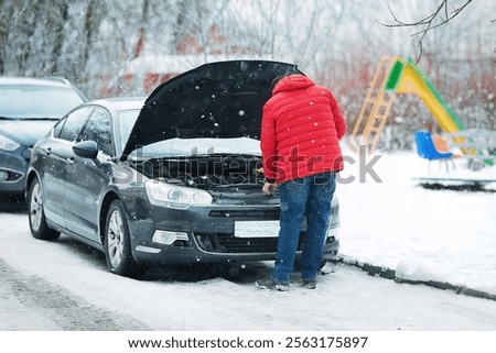 ar breakdown, Man in red jacket checking car engine under an open hood during snowfall. Vehicle troubleshooting, man inspects car engine under cold weather conditions. Fluid levels control in winter.