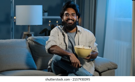 Arab Adult Bearded Man In Glasses Indian Male Bachelor After Work Sits On Sofa In Dark Living Room Turns On TV With Remote Control Choosing Channel Watching Online Movie Film Program Eating Popcorn