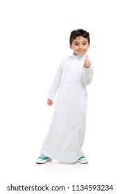 Arab boy with thumbs up and a smile, leaning to the side, wearing white traditional Saudi Thobe and sneakers, raising his hands on white isolated background