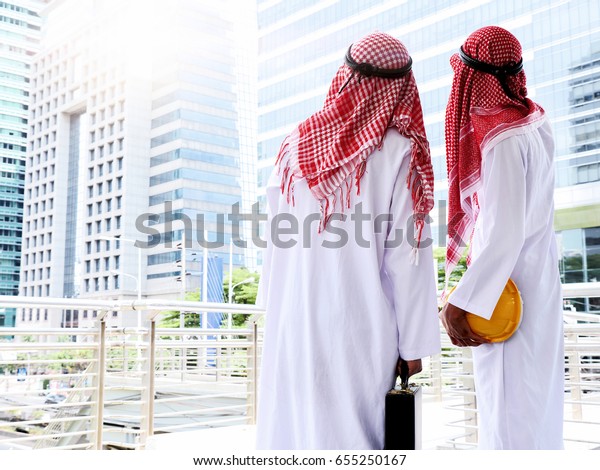 Arab businessman\
with briefcase and helmet pointing to building while working on\
project, teamwork\
concepts.