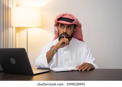 Arab Businessman In Turban Thinking While Writing At Work Desk With Office Space Background