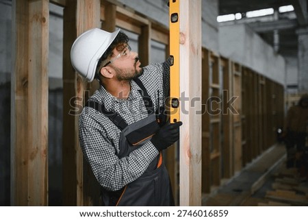 Arab construction worker using a spirit level ensuring vertical alignment of wooden beam during modular house production in a bright workshop