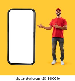 Arab Delivery Guy Showing Huge Phone Screen Pointing Fingers At Big Cellphone Advertising Online Courier Service App Over Yellow Background. Man Standing Near Phone Showing Delivering Application