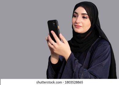 Arab Emirati Lady Using Mobile Phone Device On Gray Background Ideal For Phone Banking, Online Booking Or Ecommerce Purchase Concept In The Middle East. Arabic Woman Uses Cellphone With Copy Space