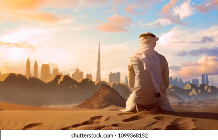 Arab Emirati Man Praying On Top Of A Dune In UAE Desert Front Dubai Skyline.