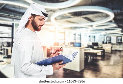 Arab Emirati man writing on a book, working in office  