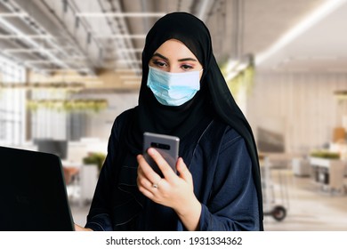 Arab With Facemask At The Office While Using Smart Mobile Phone At Work. Woman In Hijab Abaya Working Using Computer Laptop While Checking Her Cellphone