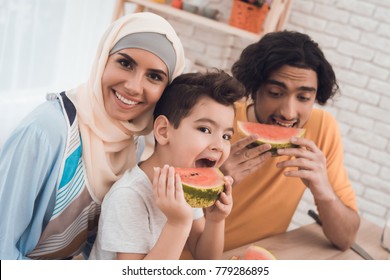The Arab family is eating a watermelon in their kitchen. A boy, a woman in a hijab and a father in modern clothes at home. Happy arabic family concept.