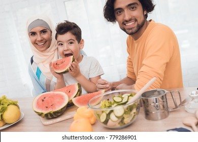 The Arab Family Is Eating A Watermelon In Their Kitchen. A Boy, A Woman In A Hijab And A Father In Modern Clothes At Home. There Are Also Other Vegetables On The Table.