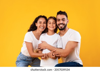 Arab Family. Happy Middle-Eastern Parents Hugging Their Kid Daughter Smiling To Camera On Yellow Background. Studio Shot Of Spouses Embracing Little Girl Expressing Their Love To Child