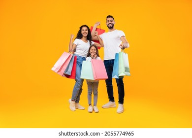 Arab Family Of Three Shopping Together Posing Holding Colorful Paper Shopper Bags Standing Over Yellow Background. Studio Shot Of Parents Ad Daughter Buying New Clothes. Full Length