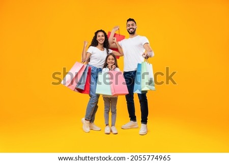 Arab Family Of Three Shopping Together Posing Holding Colorful Paper Shopper Bags Standing Over Yellow Background. Studio Shot Of Parents Ad Daughter Buying New Clothes. Full Length